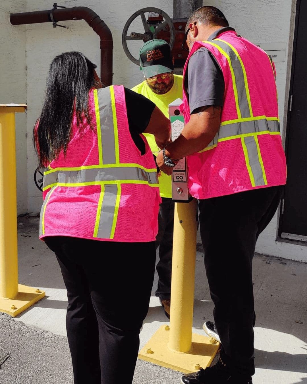 Two workers in bright pink hi-vis vests, seen from behind, fit a slim metal cigarette-butt receptacle to a black pole on a concrete pad while a third man in a green Coral Gables cap and yellow hi-vis shirt steadies it from the far side.