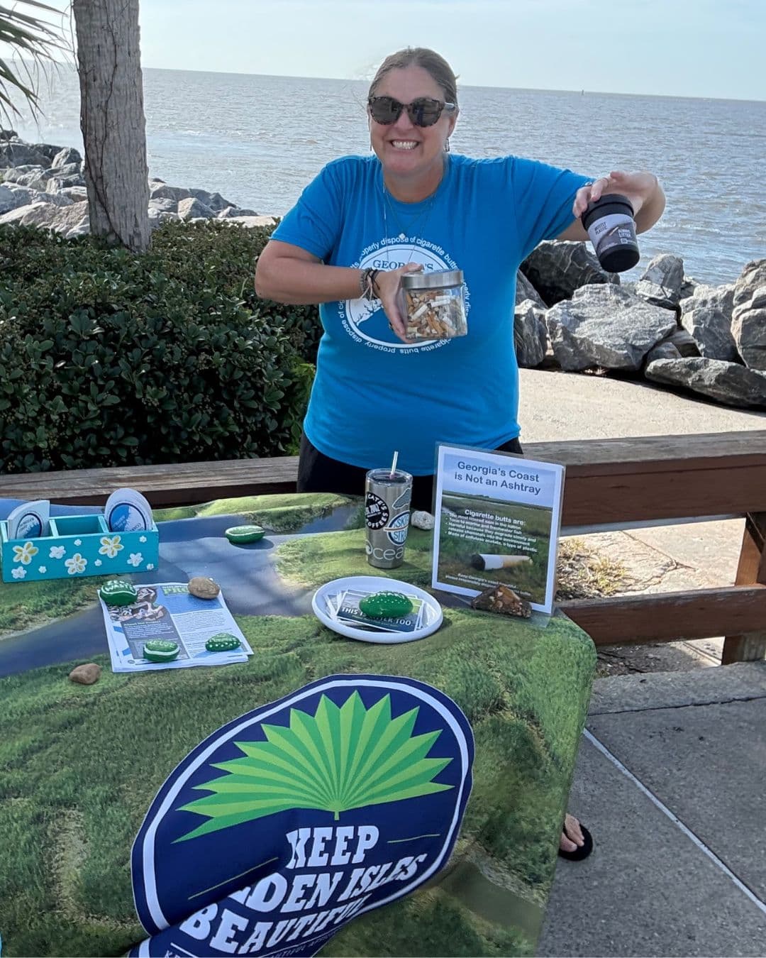 A woman in a blue t-shirt and sunglasses stands at an outdoor table by a rocky shoreline, tipping a glass jar packed with cigarette butts and holding a black pocket ashtray. A sign on the table reads Georgia's Coast is Not an Ashtray and a navy KEEP GOLDEN ISLES BEAUTIFUL banner hangs at the front.