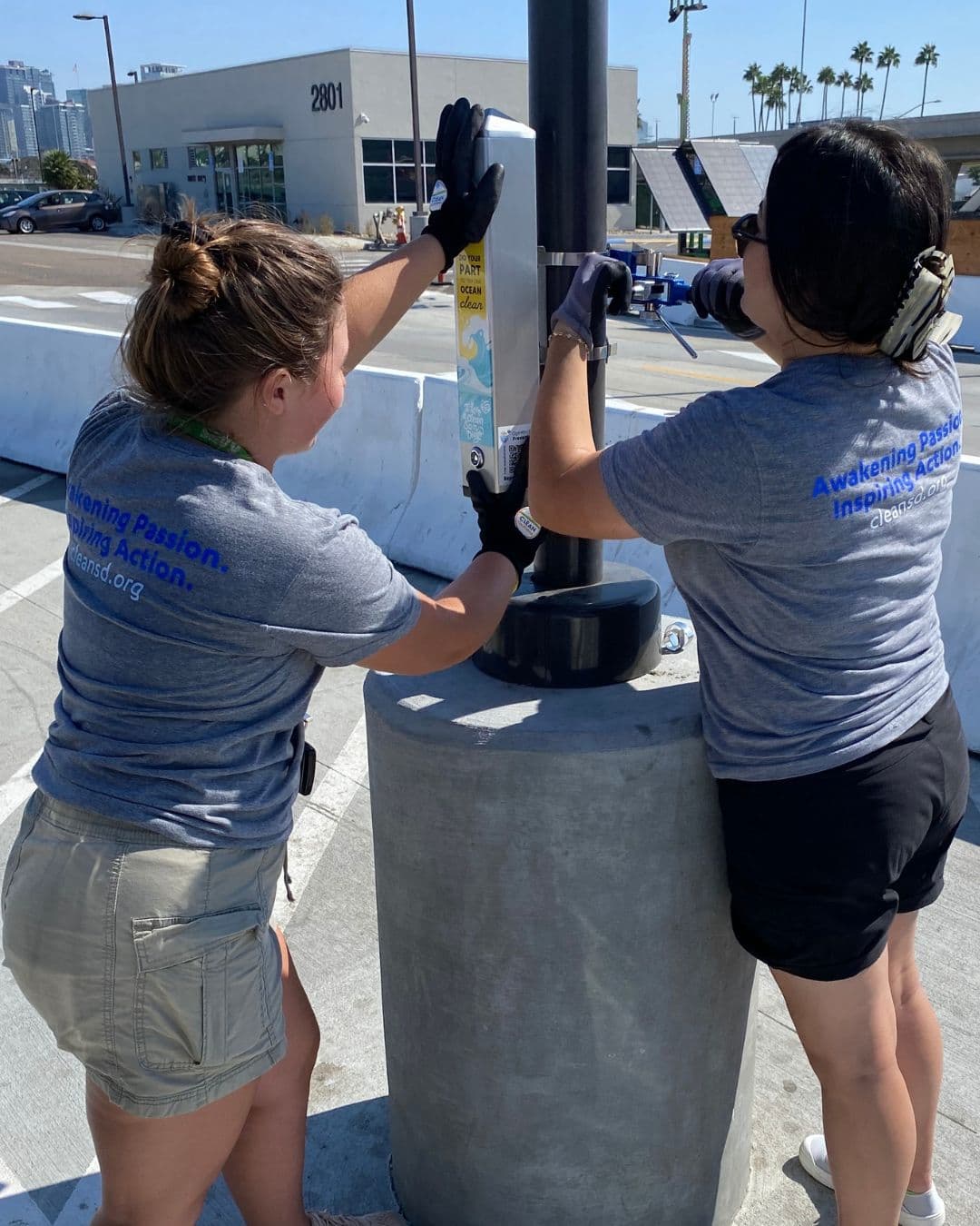 Two women in grey t-shirts printed Awakening Passion. Inspiring Action. cleansd.org bolt a white cigarette-butt receptacle to a black lamp post on a concrete waterfront kerb, palm trees and a building marked 2801 behind them.