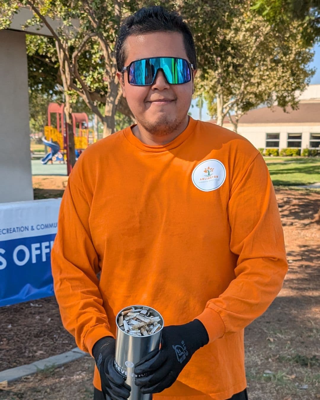 A young man in an orange long-sleeved shirt with an Arlington Community Action Group badge and wraparound mirrored sunglasses stands in a park holding up a steel canister filled to the brim with cigarette butts, black work gloves on his hands.