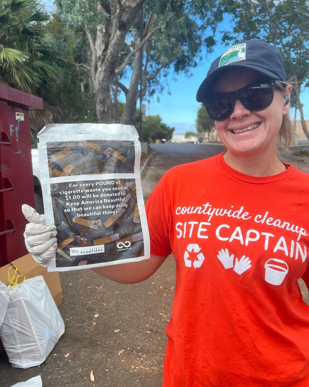 A smiling woman in a red countywide cleanup SITE CAPTAIN t-shirt, cap and sunglasses holds up a sealed pouch of cigarette waste. Text on the pouch reads that for every pound of cigarette waste sent in, one dollar goes to Keep America Beautiful.