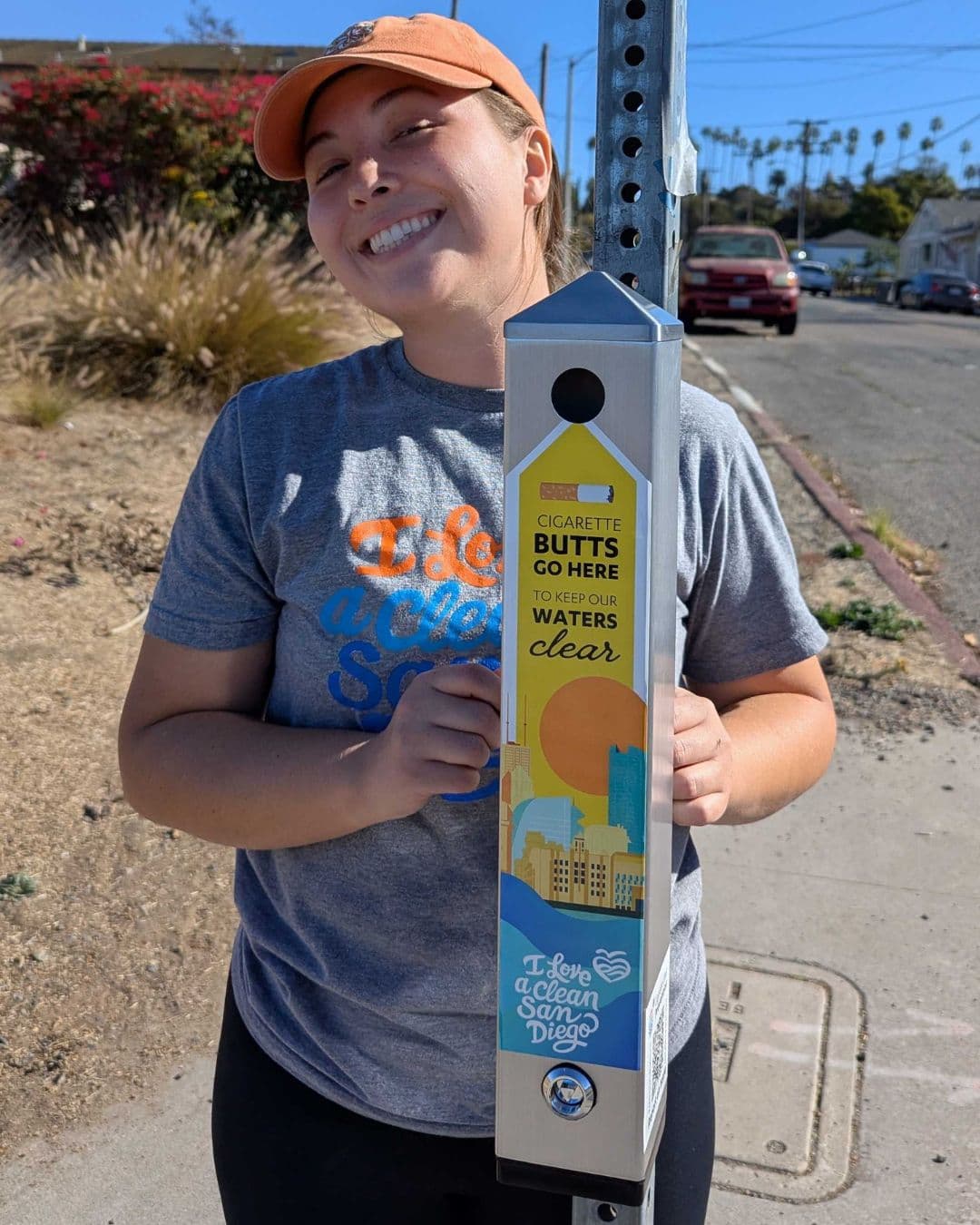 A grinning woman in an orange cap and grey t-shirt hugs a silver cigarette-butt receptacle mounted on a street pole. Its yellow decal reads CIGARETTE BUTTS GO HERE TO KEEP OUR WATERS CLEAR above an illustrated city skyline and the words I Love a Clean San Diego.