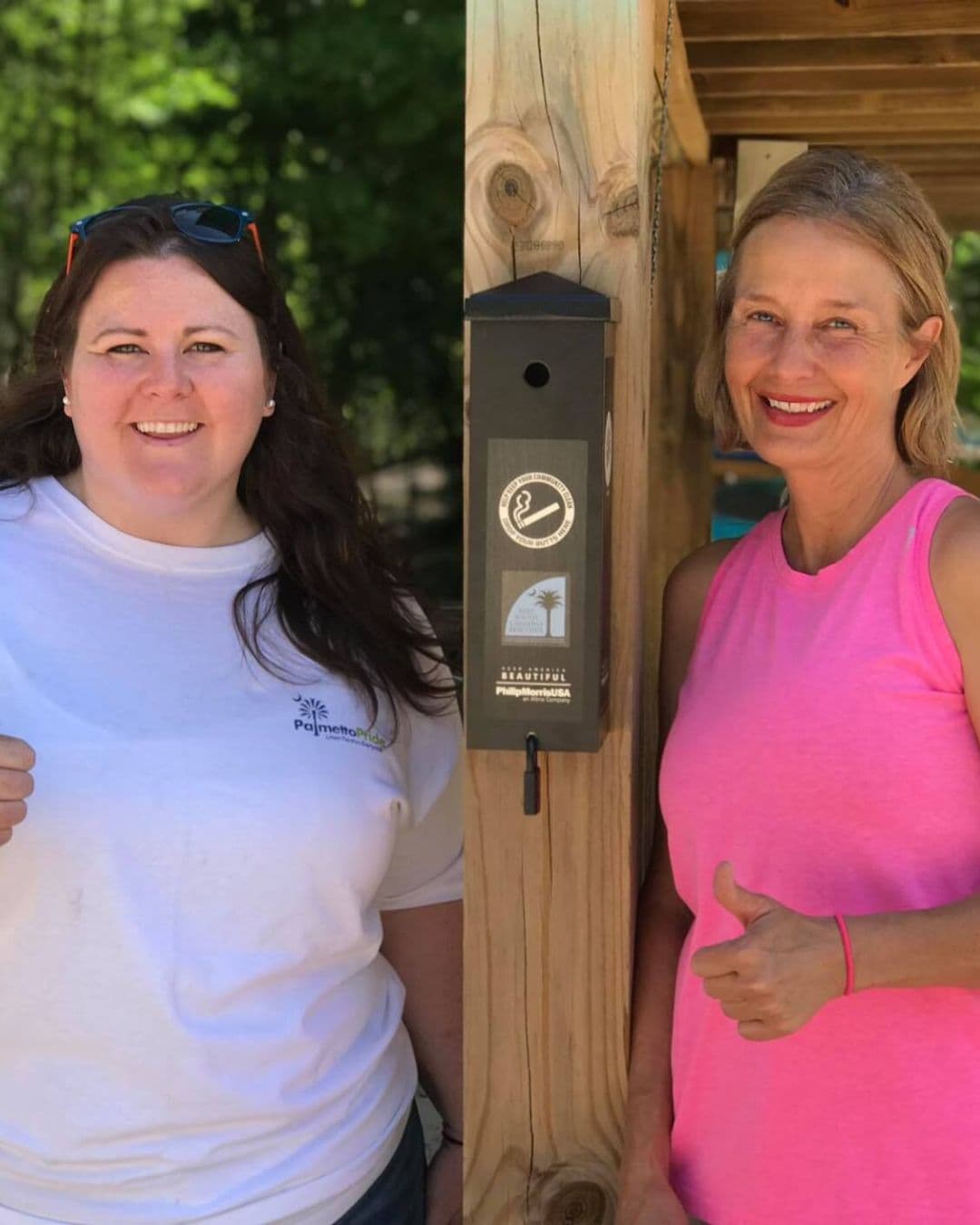 Two women stand either side of a wooden post carrying a dark grey cigarette receptacle labelled with a crossed-cigarette symbol, a palmetto badge, KEEP AMERICA BEAUTIFUL and Philip Morris USA. The woman on the left wears a white PalmettoPride t-shirt; the woman on the right in a pink vest gives a thumbs up.