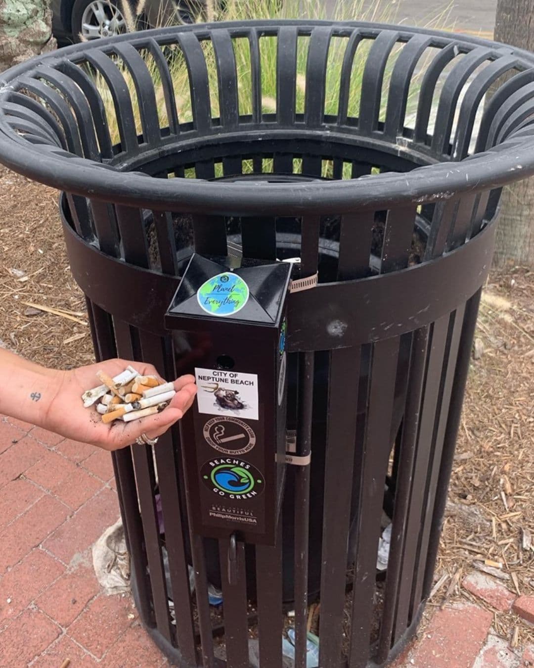 An outstretched hand holds a pile of spent cigarette butts in front of a black slatted street bin. A narrow ashtray attached to the bin bears a City of Neptune Beach card, a BEACHES GO GREEN roundel and an Earth sticker reading Planet Everything.