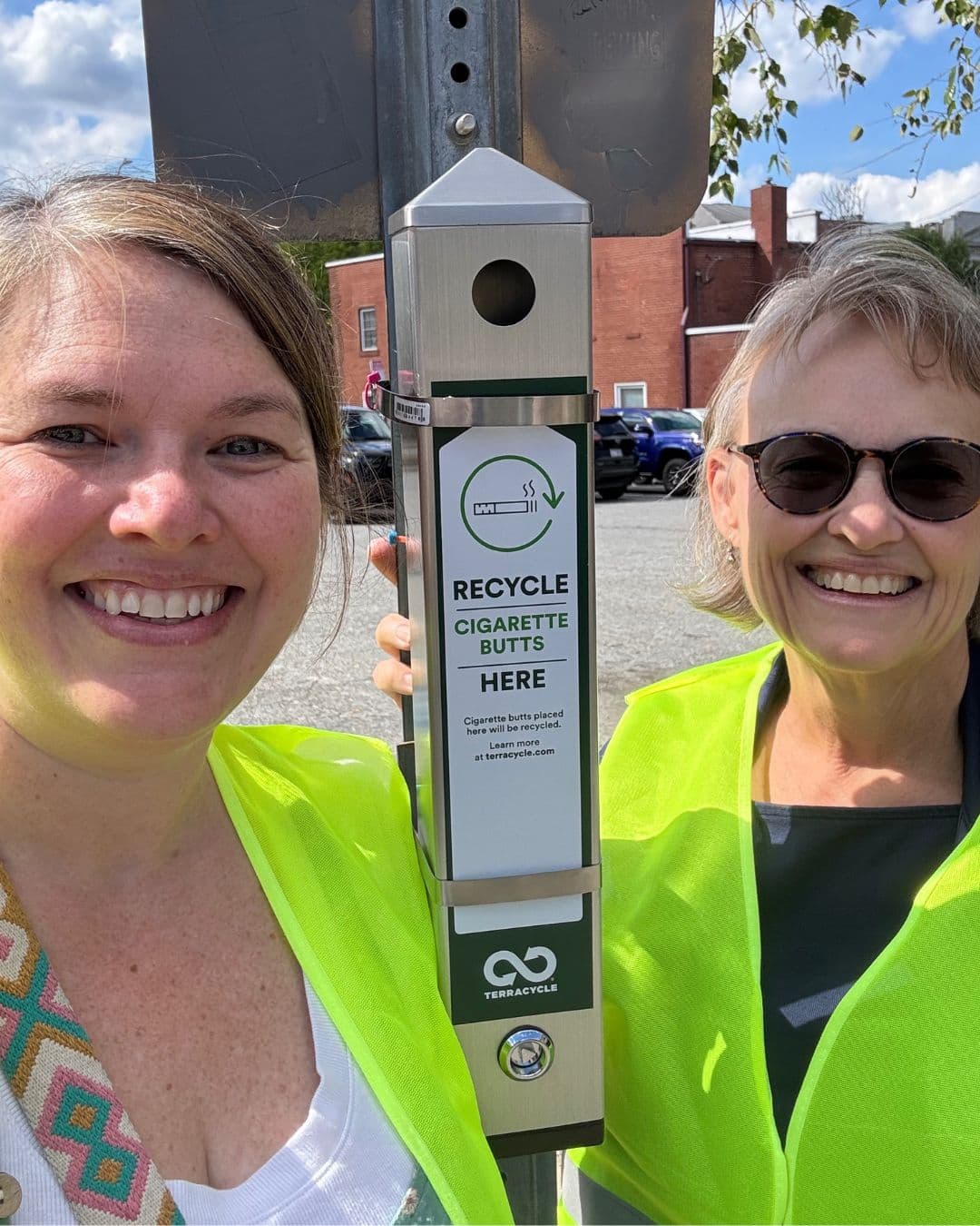 Close selfie of two smiling women in yellow hi-vis vests either side of a steel post-mounted receptacle. Its panel reads RECYCLE CIGARETTE BUTTS HERE, with a note that butts placed there will be recycled and a TerraCycle logo below.