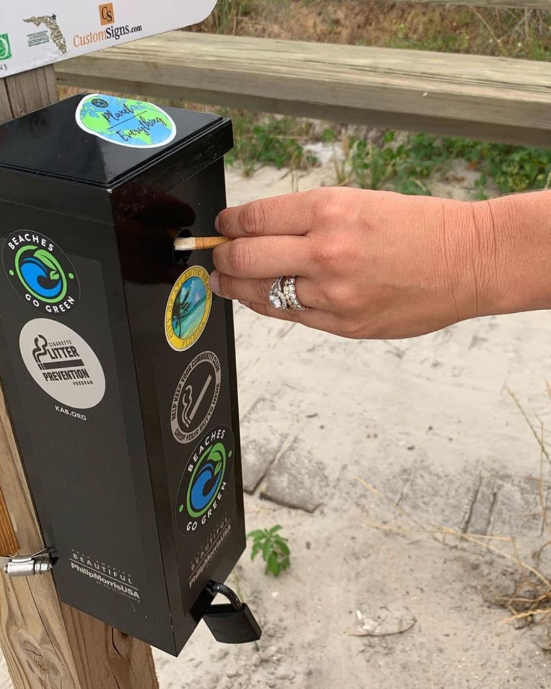 A ringed hand pushes a cigarette into the slot of a black post-mounted ashtray on a sandy boardwalk. Stickers on the box read BEACHES GO GREEN, CIGARETTE LITTER PREVENTION PROGRAM KAB.ORG, and Keep America Beautiful with Philip Morris USA.