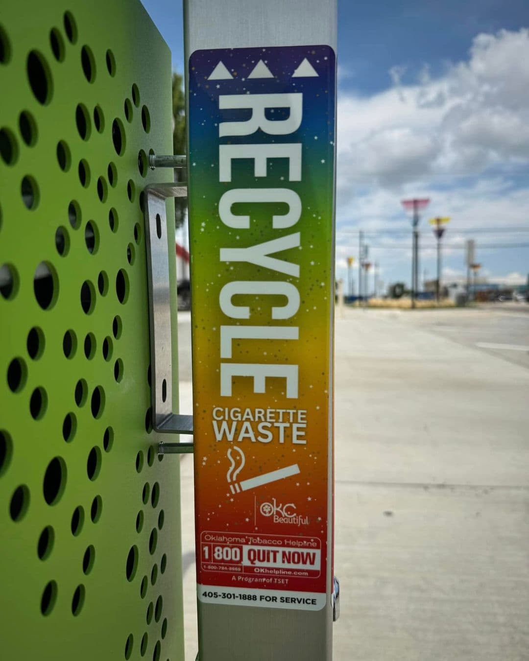 Close view of a rainbow-gradient decal on a steel post receptacle standing beside a lime-green perforated bin in a sunlit car park. The lettering reads RECYCLE CIGARETTE WASTE with an OKC Beautiful mark and an Oklahoma Tobacco Helpline 1-800-QUIT-NOW panel.
