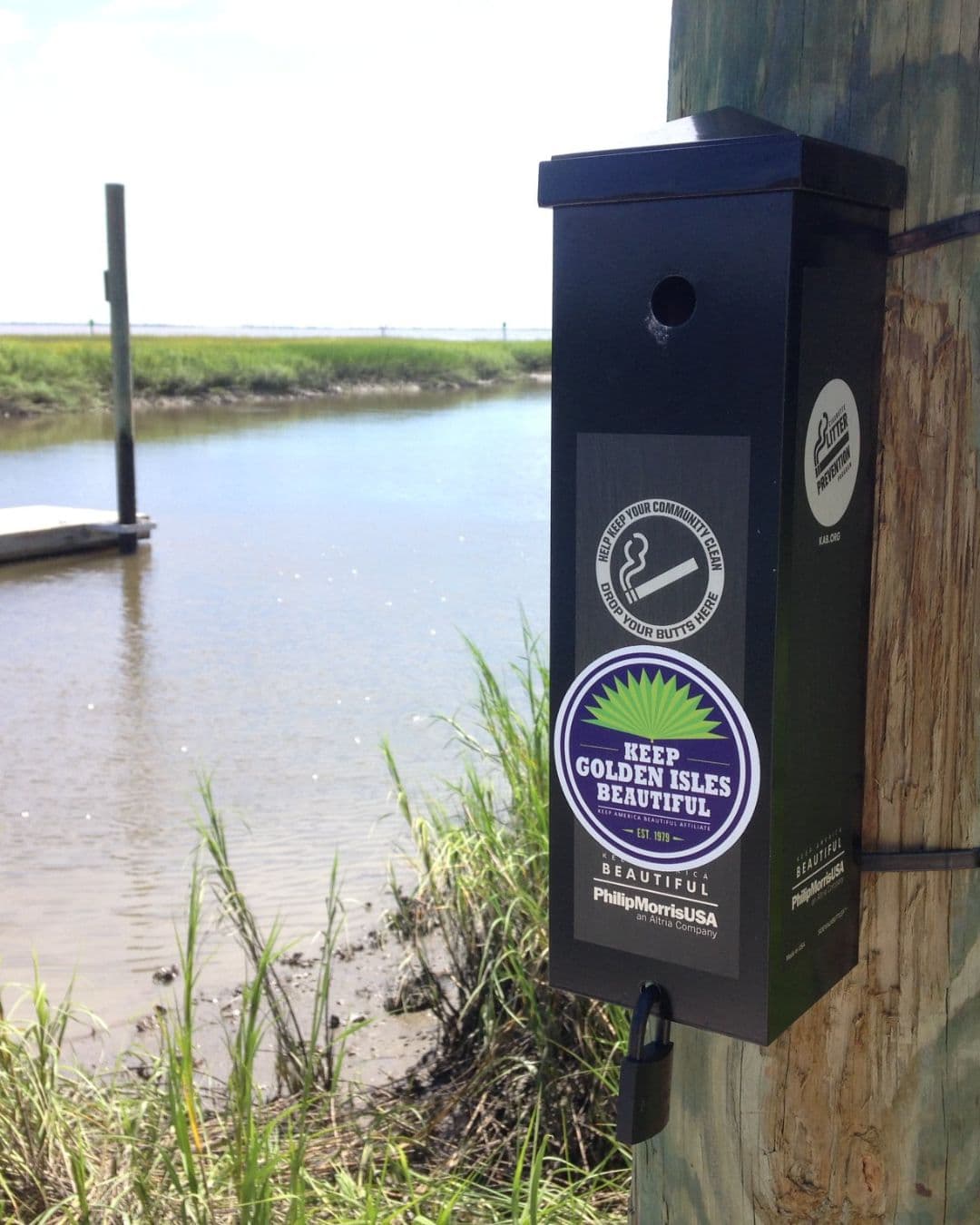 A black padlocked cigarette receptacle strapped to a weathered wooden piling above a tidal marsh creek. Its decals read HELP KEEP YOUR COMMUNITY CLEAN DROP YOUR BUTTS HERE and a purple KEEP GOLDEN ISLES BEAUTIFUL roundel, with Philip Morris USA beneath.