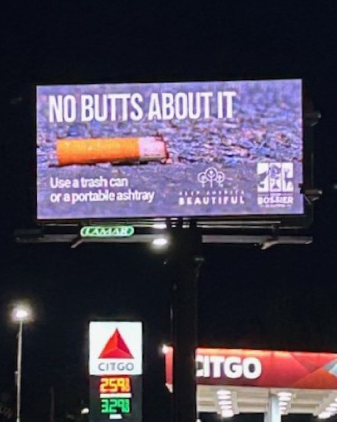 A lit roadside billboard at night showing a discarded cigarette on asphalt with the headline NO BUTTS ABOUT IT and the line Use a trash can or a portable ashtray. A Citgo petrol station with fuel prices glows below.