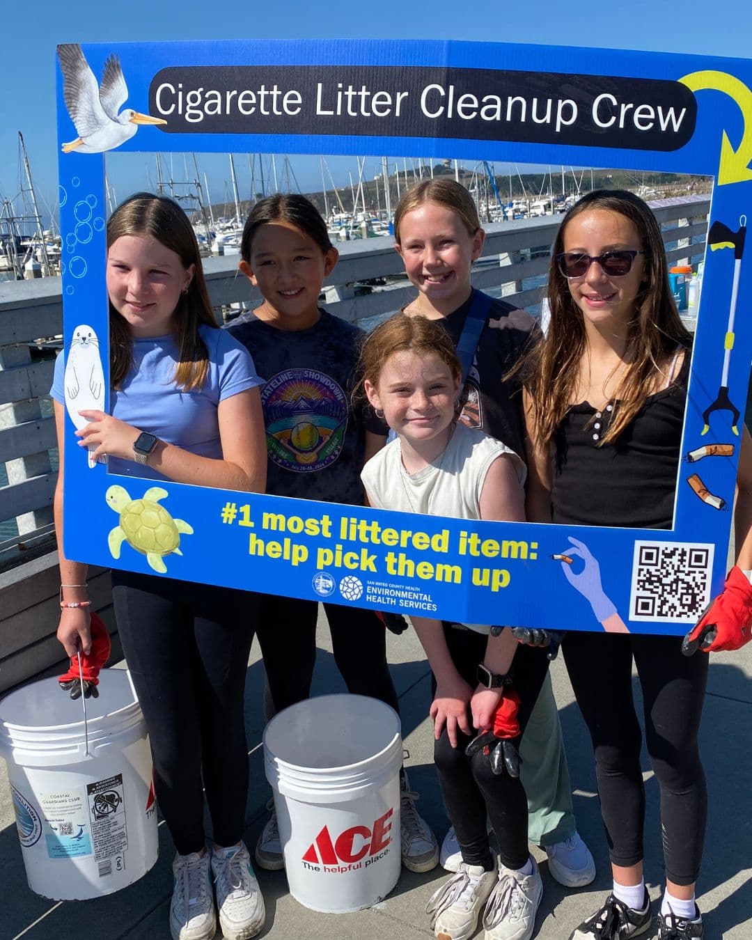 Five girls stand on a harbour pier holding a large blue photo frame titled Cigarette Litter Cleanup Crew, its lower edge reading number one most littered item: help pick them up. Two white buckets and red work gloves are at their feet, moored sailboats behind.
