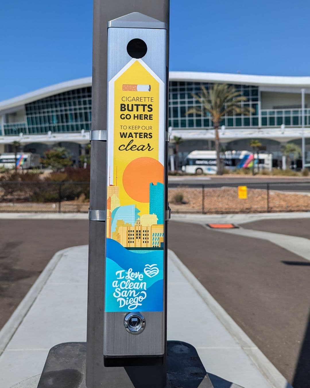 A brushed-steel cigarette receptacle clamped to a grey pole on a sunny plaza, its decal reading CIGARETTE BUTTS GO HERE TO KEEP OUR WATERS CLEAR over an illustrated skyline and the I Love a Clean San Diego mark. A long curved glass building and palms stand behind.