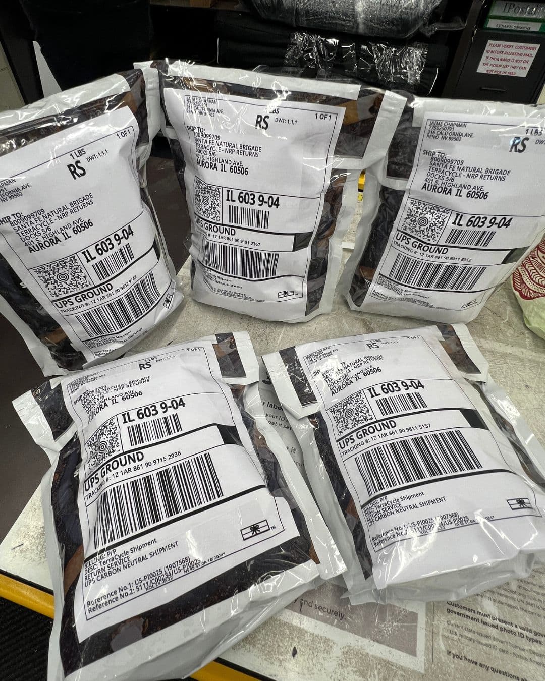 Five foil pouches of dark cigarette waste laid out on a shop counter, each carrying a large UPS Ground shipping label addressed to Santa Fe Natural Brigade, TerraCycle NRP Returns in Aurora, Illinois.