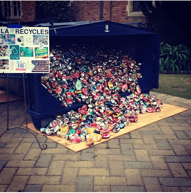 A dark blue skip-style container on a brick courtyard, its open side spilling hundreds of crushed aluminium drink cans onto a plywood sheet. A display board on an easel to the left reads RECYCLES with recycling diagrams.
