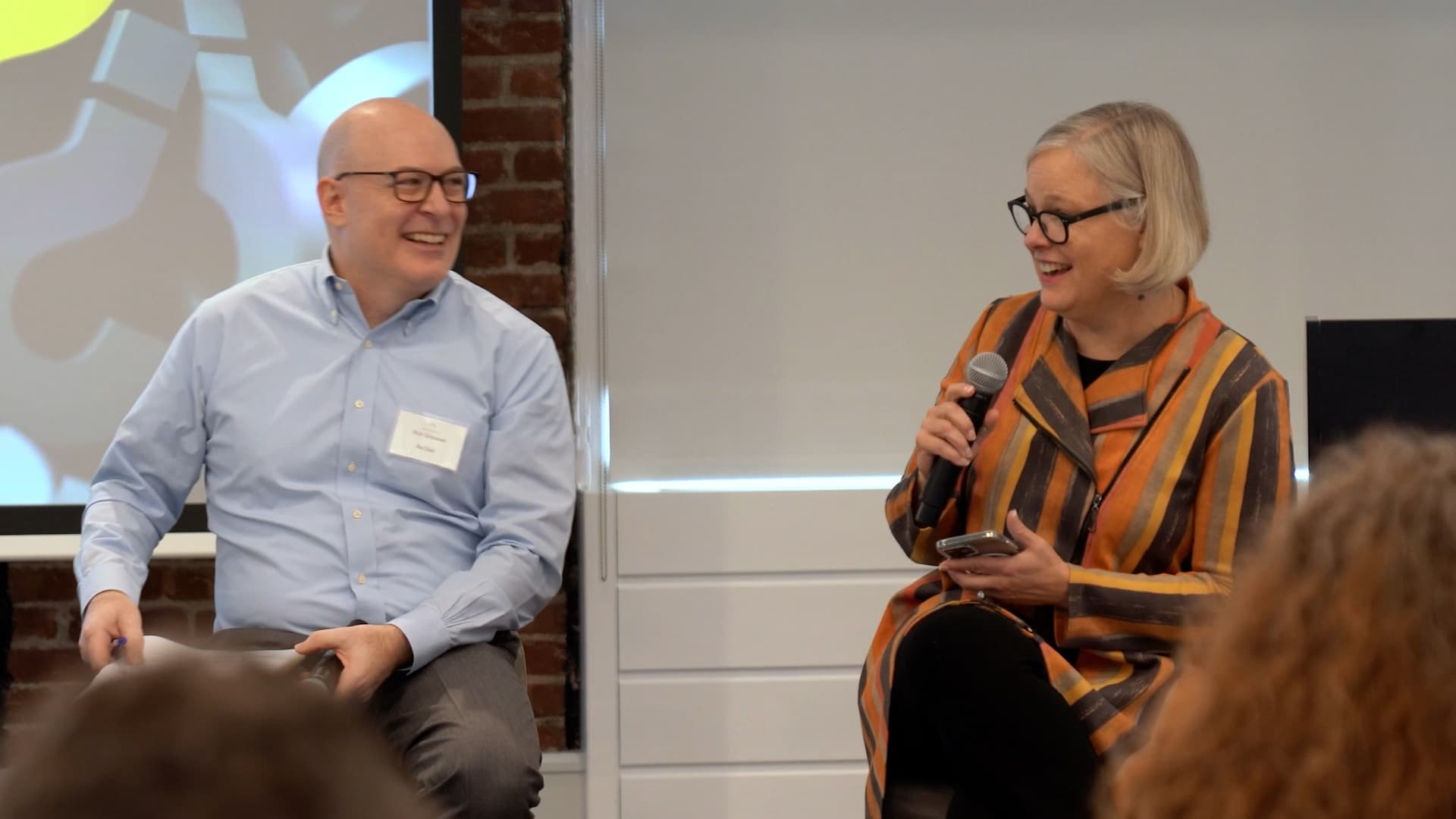 A man in a pale blue shirt, glasses and a name badge sits smiling on a low stage beside a woman with a blonde bob in a striped orange and brown coat who is speaking into a handheld microphone. A projection screen and exposed brick wall are behind them, audience heads in the foreground.