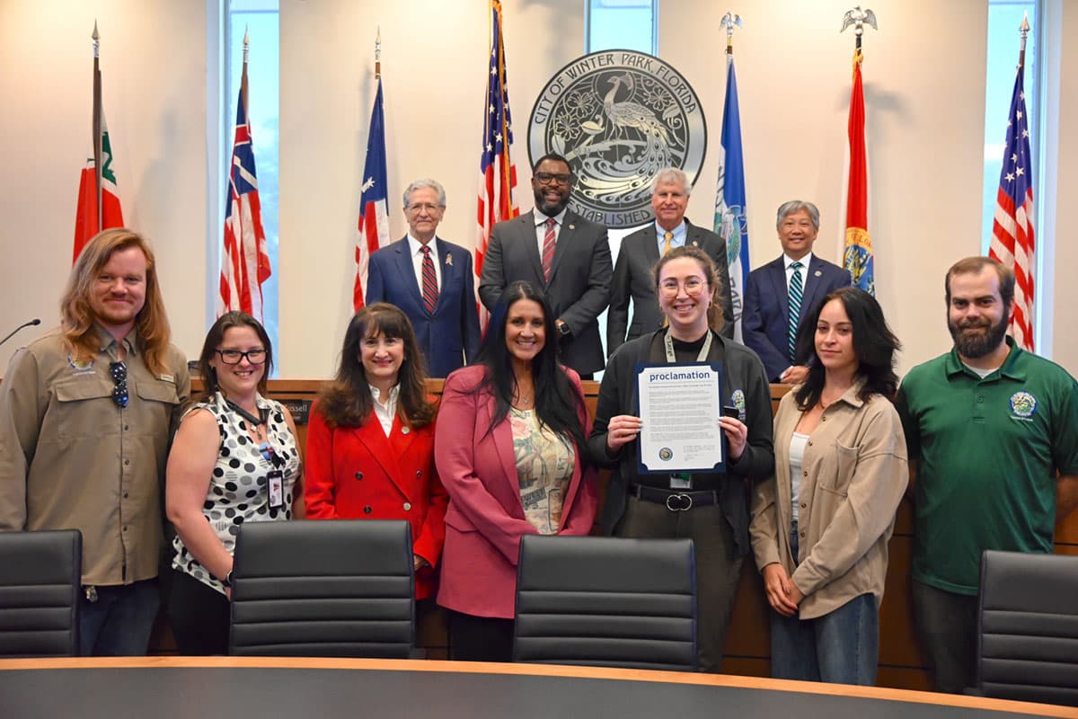 A group of about a dozen people pose in a council chamber in front of a wall of flags and a seal reading CITY OF WINTER PARK FLORIDA. A woman in the centre front holds open a navy folder headed proclamation; four men in suits stand behind the dais.