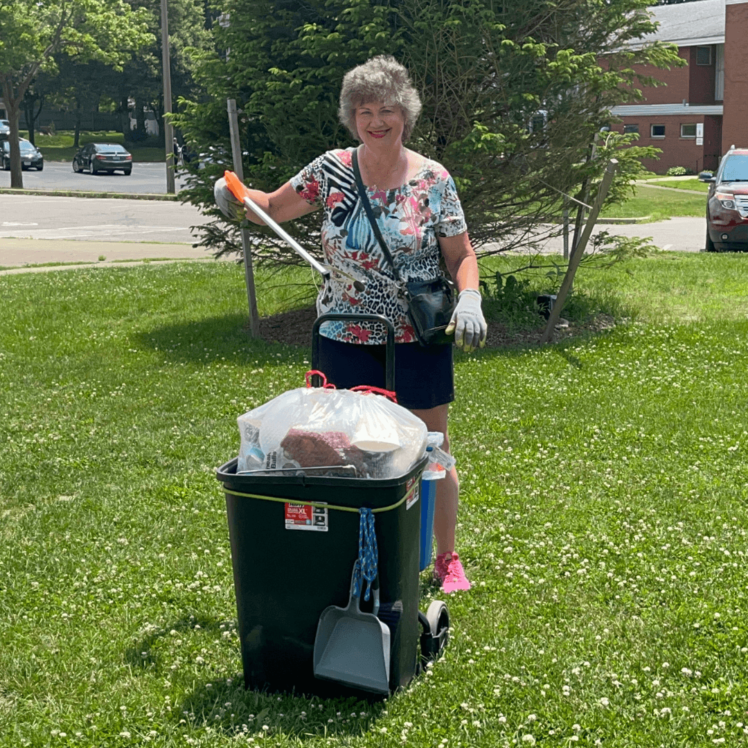 Woman in a floral top holds a litter grabber beside a green wheeled bin topped with a bagged haul and a dustpan on a sunny lawn