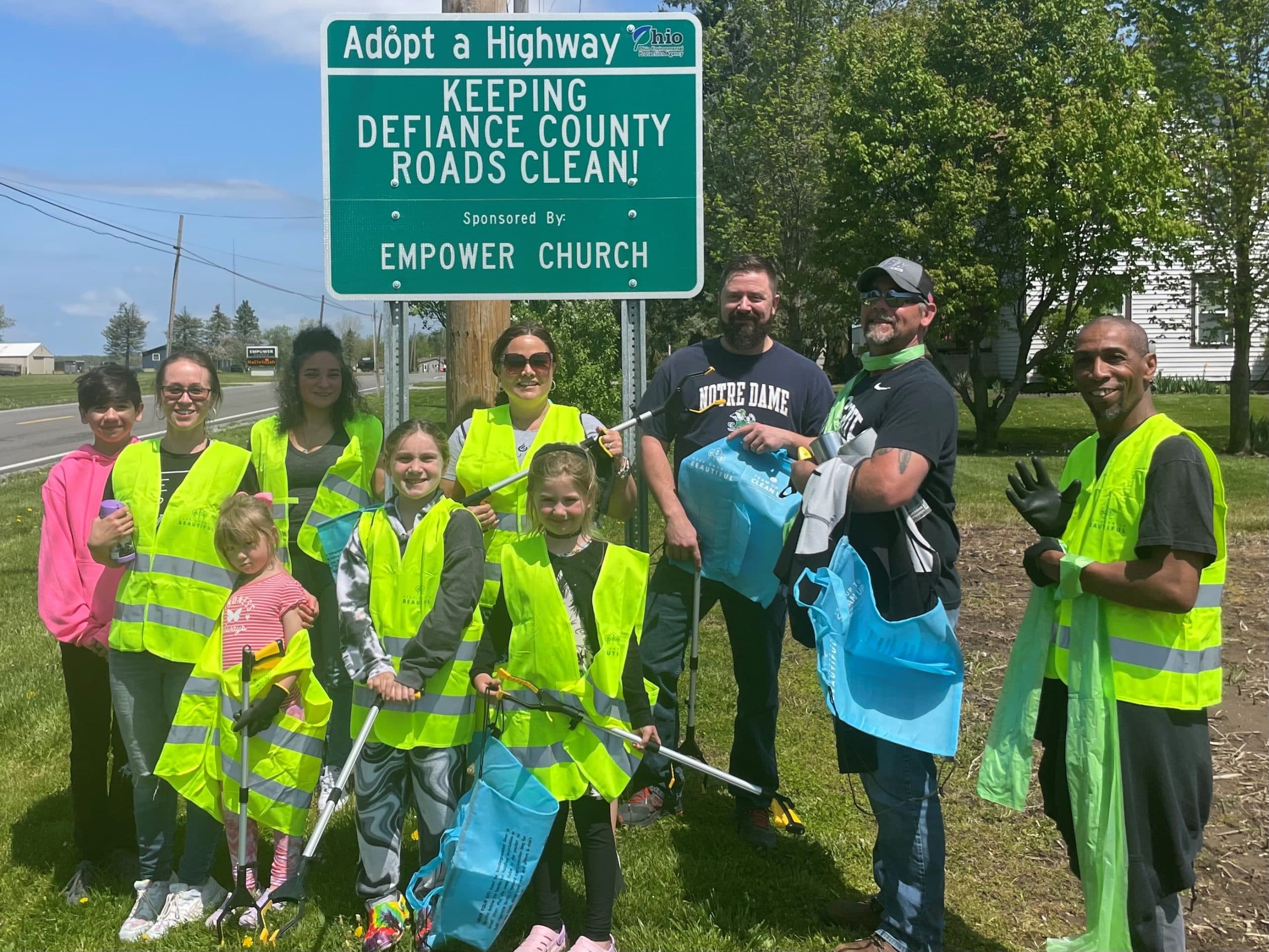 Families in lime vests with grabbers and blue tote bags beneath an Adopt a Highway sign reading KEEPING DEFIANCE COUNTY ROADS CLEAN!