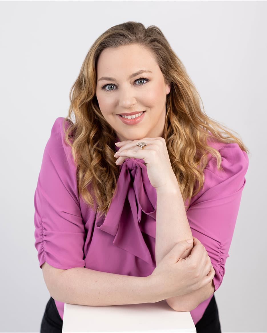 Studio portrait of a woman with long wavy blonde hair in a magenta blouse with a tie neck, resting her chin on one hand and leaning on a white plinth against a pale grey background.