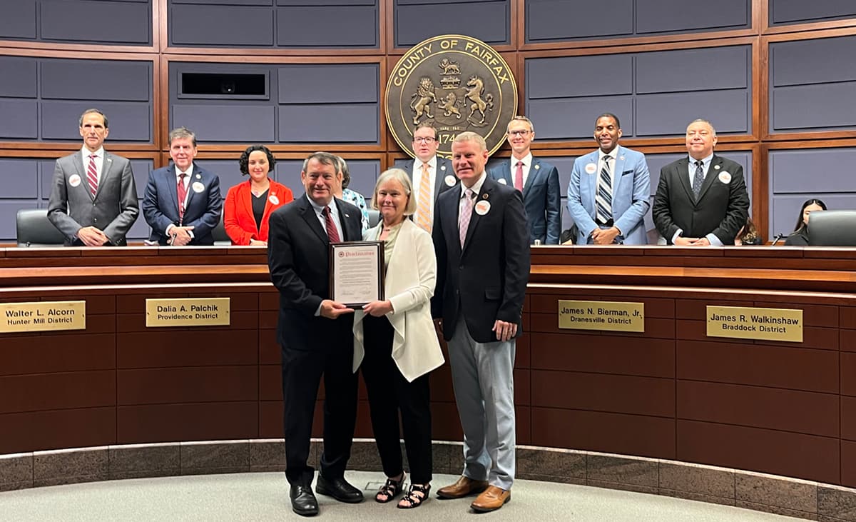 Three people stand in a county board chamber holding a framed proclamation between them, with eight officials lined up behind the curved dais under a bronze COUNTY OF FAIRFAX seal. Desk nameplates read Walter L. Alcorn, Dalia A. Palchik, James N. Bierman, Jr. and James R. Walkinshaw.