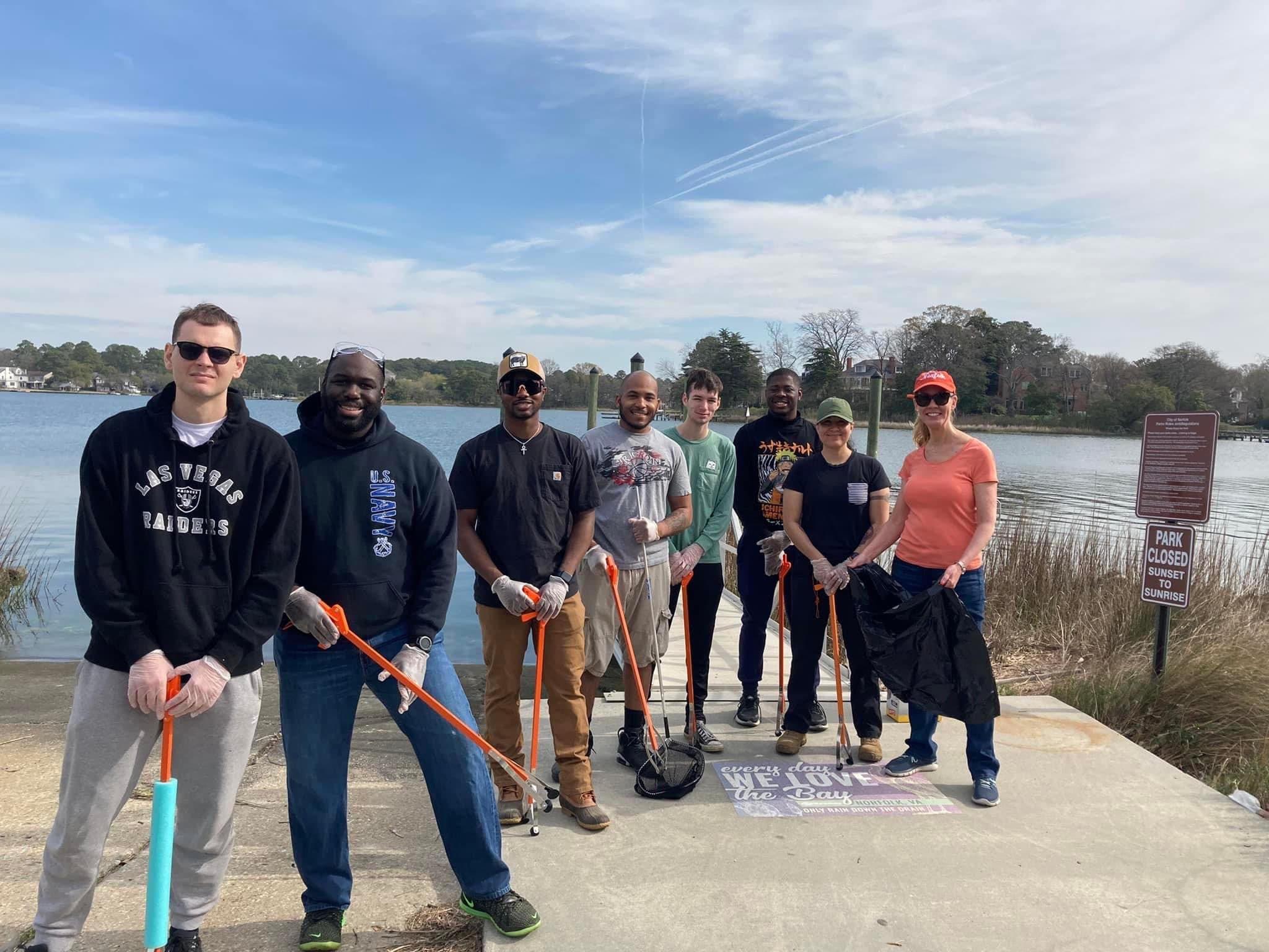 Small view of eight volunteers with orange grabbers standing on a concrete waterfront pier, a painted mural at their feet