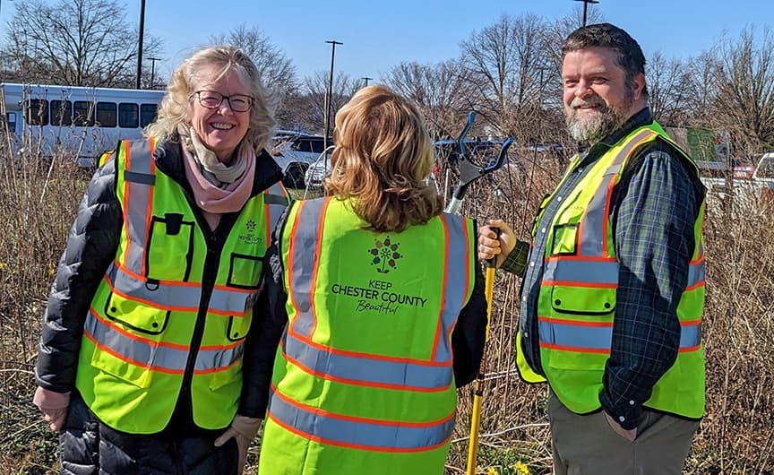 Three volunteers in lime hi-vis vests, one lettered KEEP CHESTER COUNTY BEAUTIFUL on the back, stand in a brushy lot near a parked bus