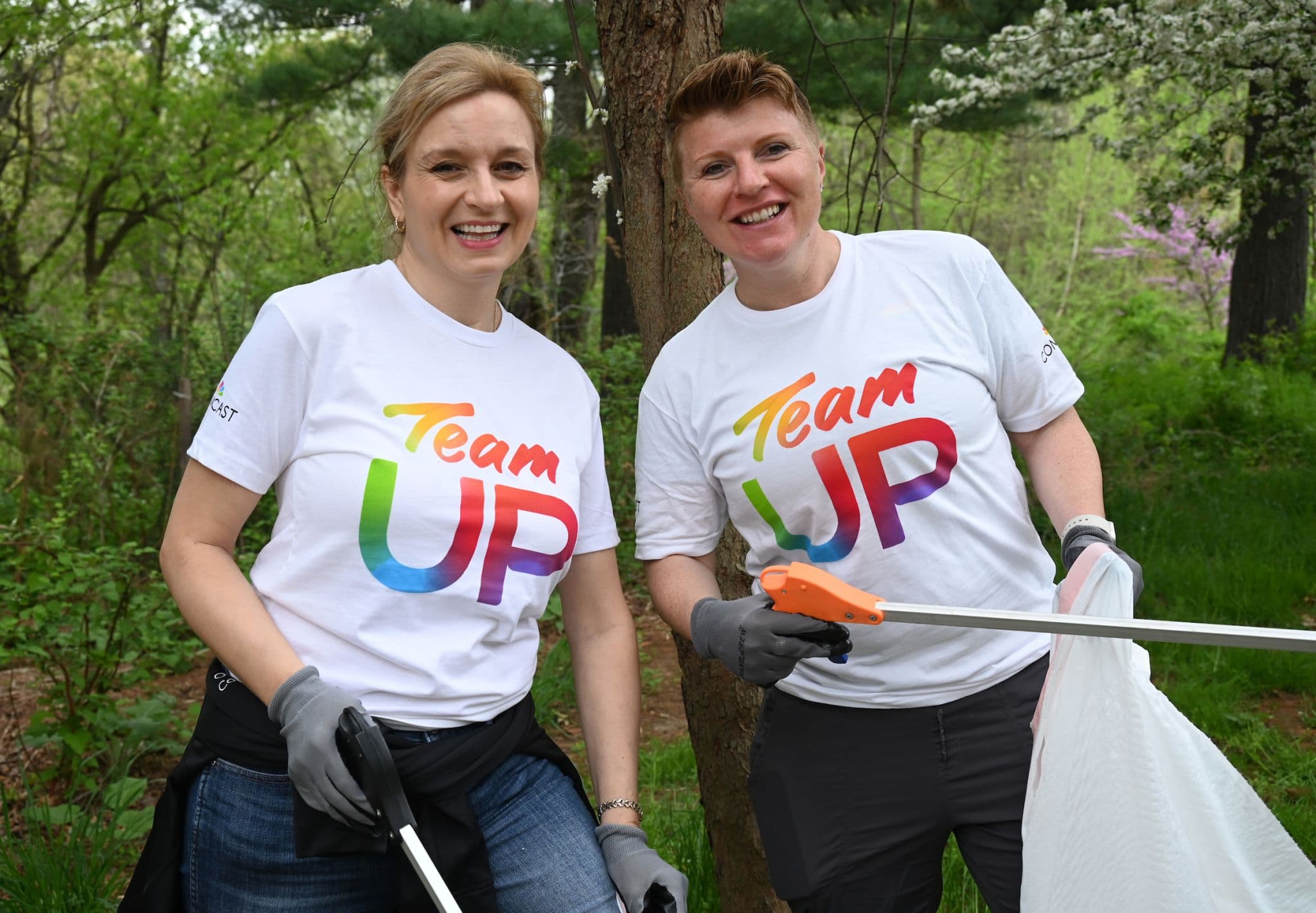 Small view of two smiling volunteers in Team UP shirts among flowering trees, one holding a grabber and a clear bag
