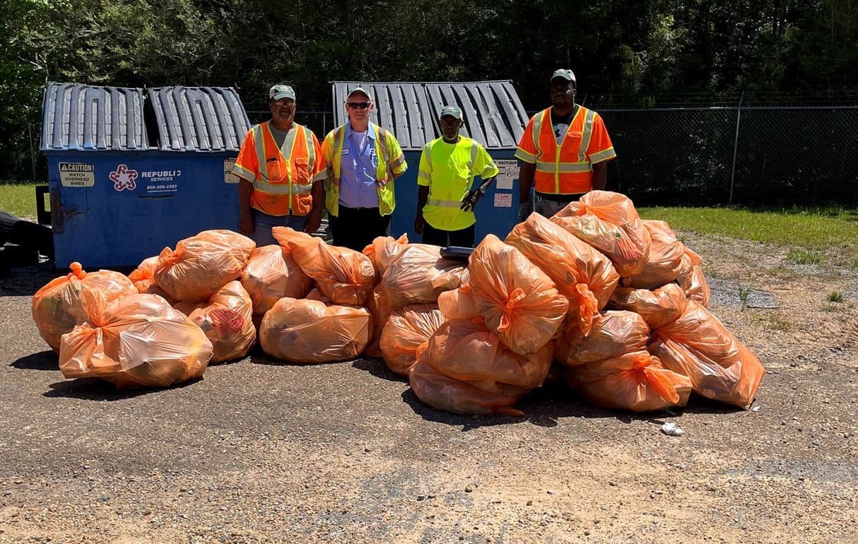 Small view of four workers standing behind a large heap of orange trash bags beside a blue dumpster
