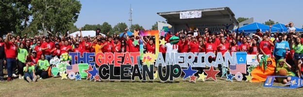 A very large crowd in red, green and blue t-shirts stands on a grass field cheering with arms raised behind giant painted cut-out letters spelling THE GREAT AMERICAN CLEAN UP, decorated with stars and flags, a covered stage behind them.