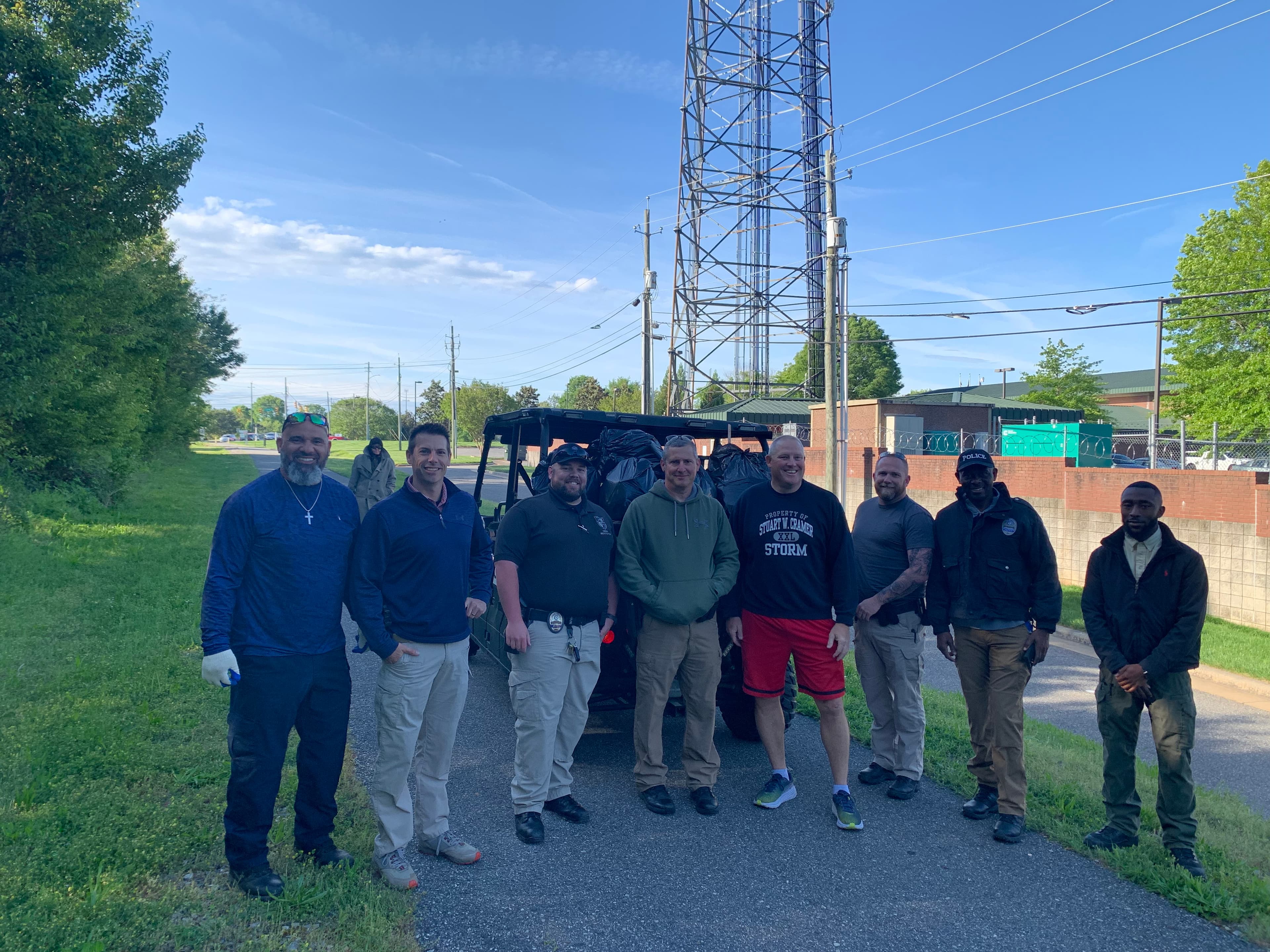 Small view of nine men standing on a paved path in front of a black utility vehicle loaded with trash bags under a lattice radio tower
