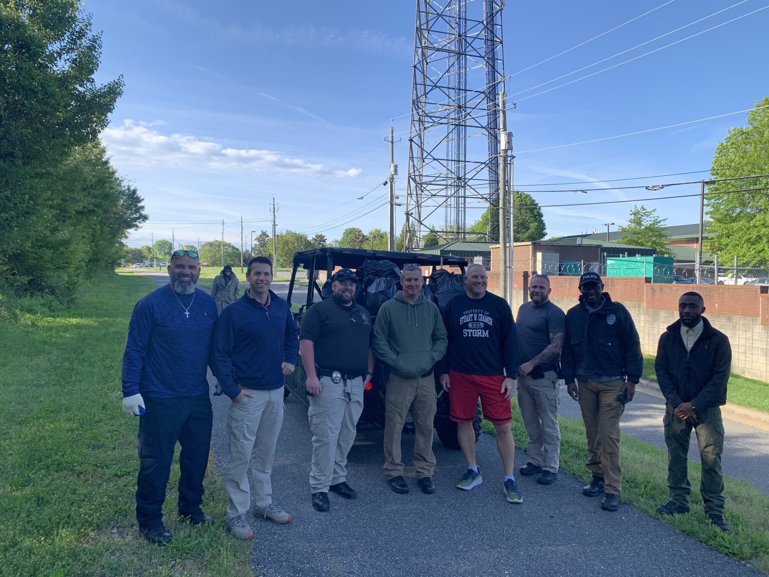 Nine officers and staff in polos and hoodies pose on a greenway path beside a utility cart piled with black trash bags, radio tower behind