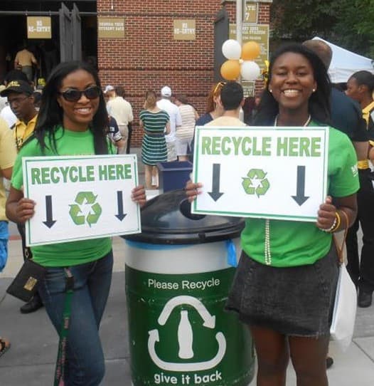 Two young women in green t-shirts stand outside a brick stadium either side of a green-and-grey bin, each holding a white sign reading RECYCLE HERE with a recycling symbol between two black arrows. The bin is printed Please Recycle and give it back around a bottle graphic.