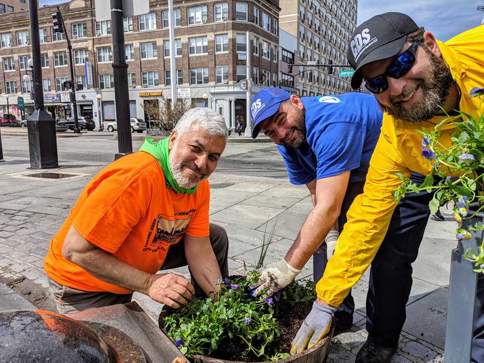 Three men crouch around a large barrel planter on a city pavement, pressing purple pansies into the soil. One wears an orange Jersey City t-shirt, one a blue polo and cap, and the bearded man on the right a yellow jacket, sunglasses and a dark cap; a brick apartment block and shopfronts stand behind.