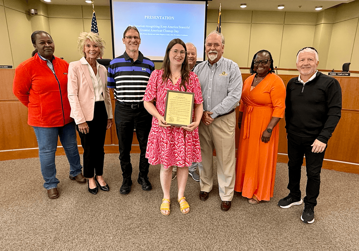 Eight people stand in a council chamber for a group photo; a woman in a pink and white patterned dress holds a framed proclamation at the centre. The screen behind reads PRESENTATION, Proclamation recognizing Keep America Beautiful Greatest American Cleanup Day, and desk nameplates are visible either side.