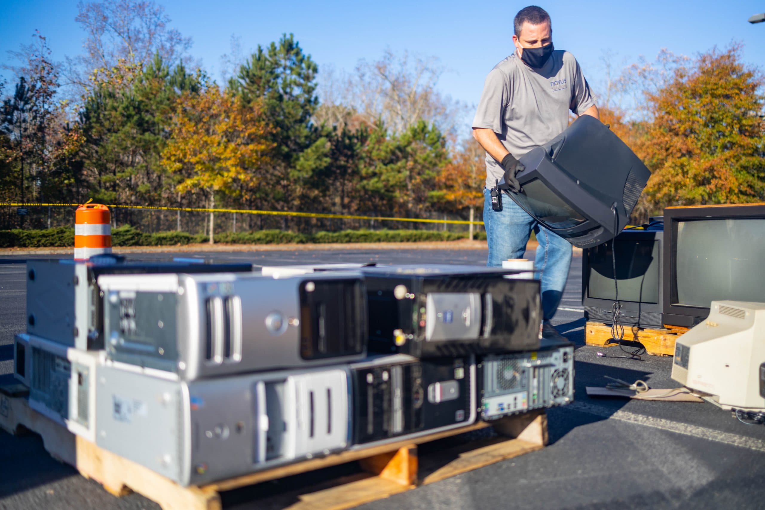 A masked man in a grey work shirt and black gloves lifts an old cathode-ray television onto a stack at an outdoor collection point in a car park, with pallets of desktop computer towers in the foreground and more televisions on pallets beside him.