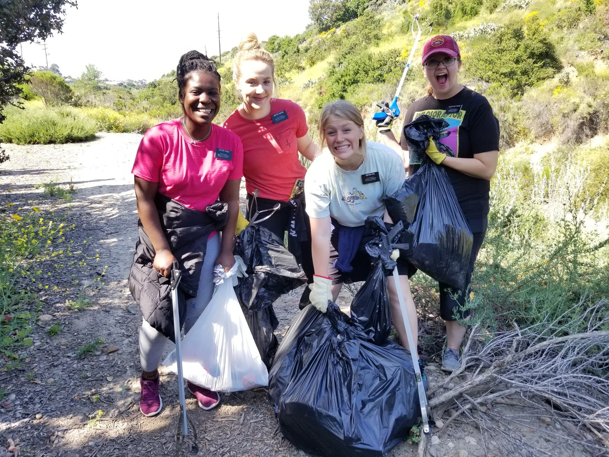 Four young women grin on a dry creekside trail holding black and white trash bags and litter grabbers, one raising a grabber overhead