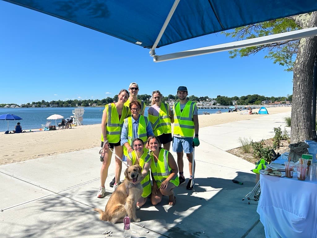 Seven volunteers in lime vests and a golden retriever pose under a blue canopy on a beach boardwalk with a lifeguard chair behind