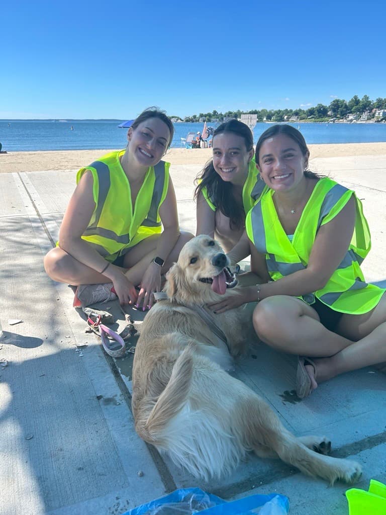 Three young women in lime safety vests sit cross-legged on a beach sidewalk petting a panting golden retriever, the bay behind them