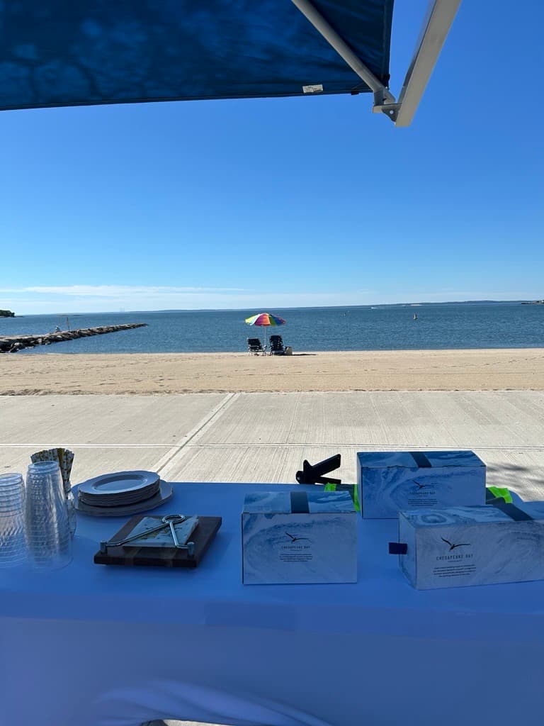 White-clothed table with stacked cups, plates and three Chesapeake Bay gift boxes under a blue canopy overlooking a beach and rainbow umbrella
