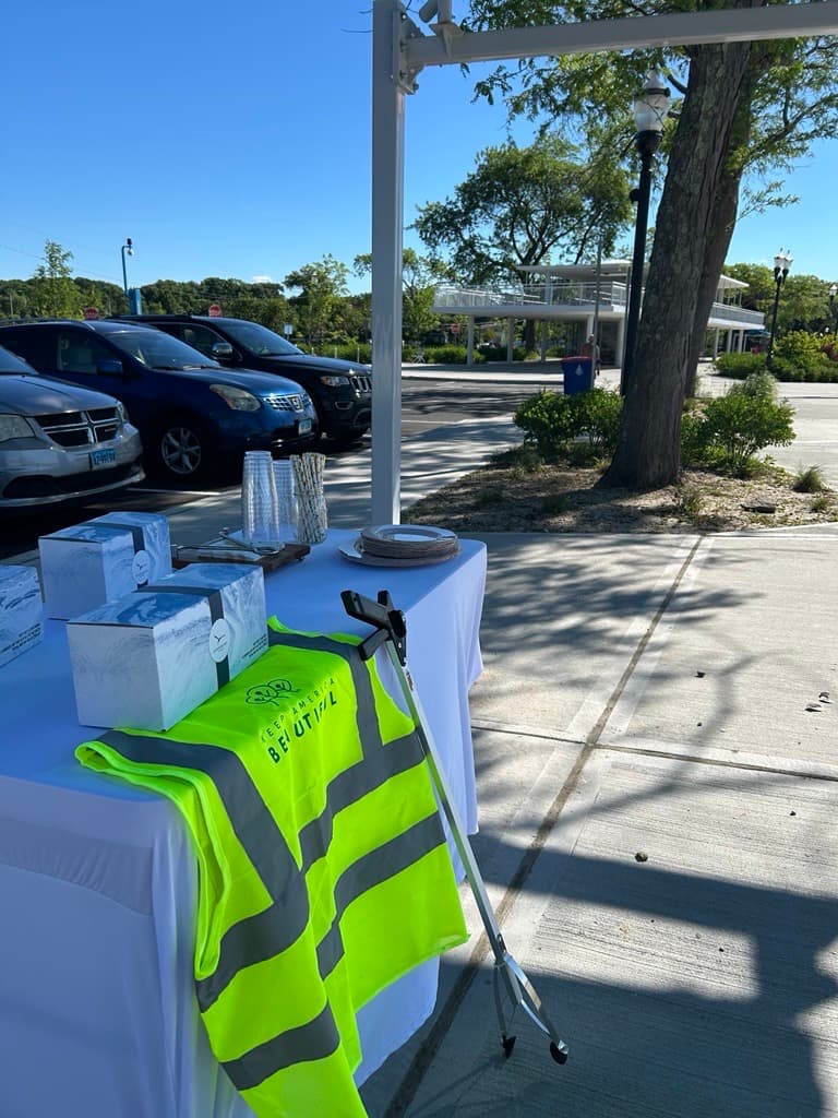 Lime Keep America Beautiful safety vest and a litter grabber draped over a white event table with gift boxes, parked cars beyond