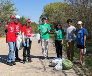 Seven teenagers in red, green and blue shirts with blue gloves stand along a gravel roadside beside filled white and green trash bags