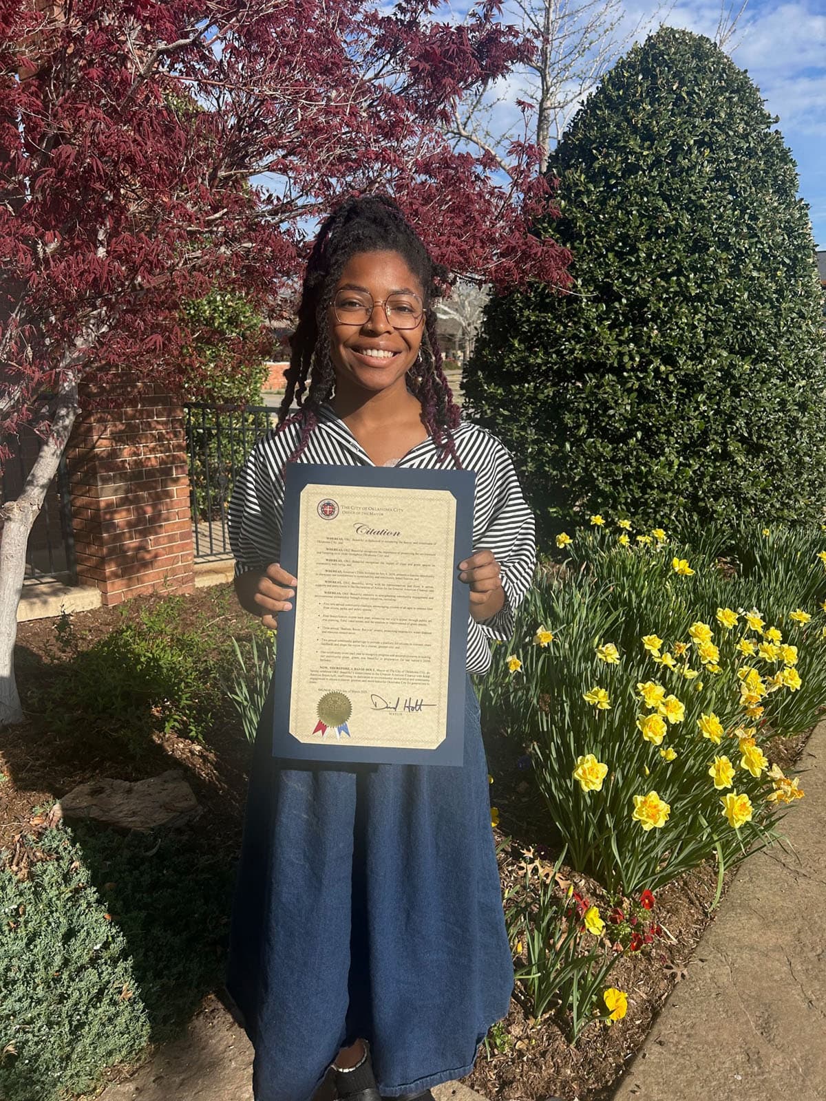 Young woman in a striped top and denim skirt holds a City of Oklahoma City mayoral Citation in front of daffodils and a red maple