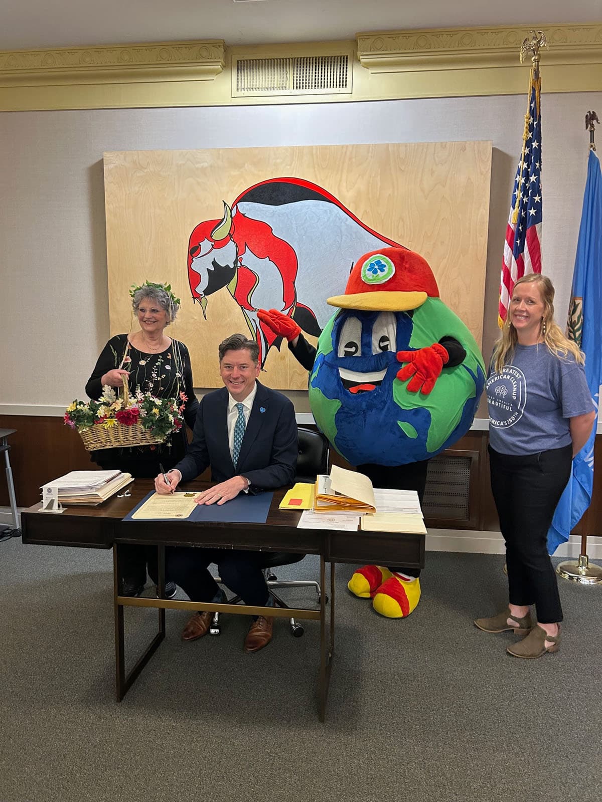 Mayor in a navy suit signs a proclamation while a woman with a flower basket, a globe mascot in a red cap and a woman in a cleanup T-shirt look on