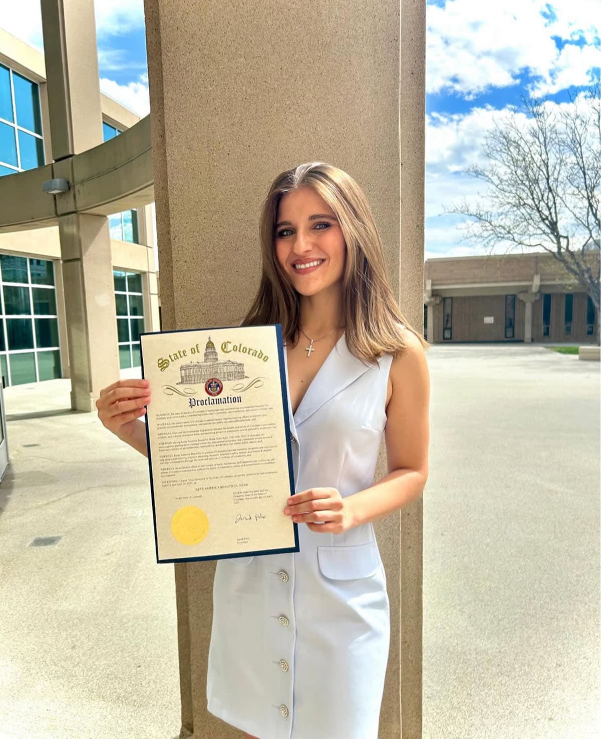 Young woman in a white dress holds a framed State of Colorado Proclamation for Keep America Beautiful Week outside a concrete civic building