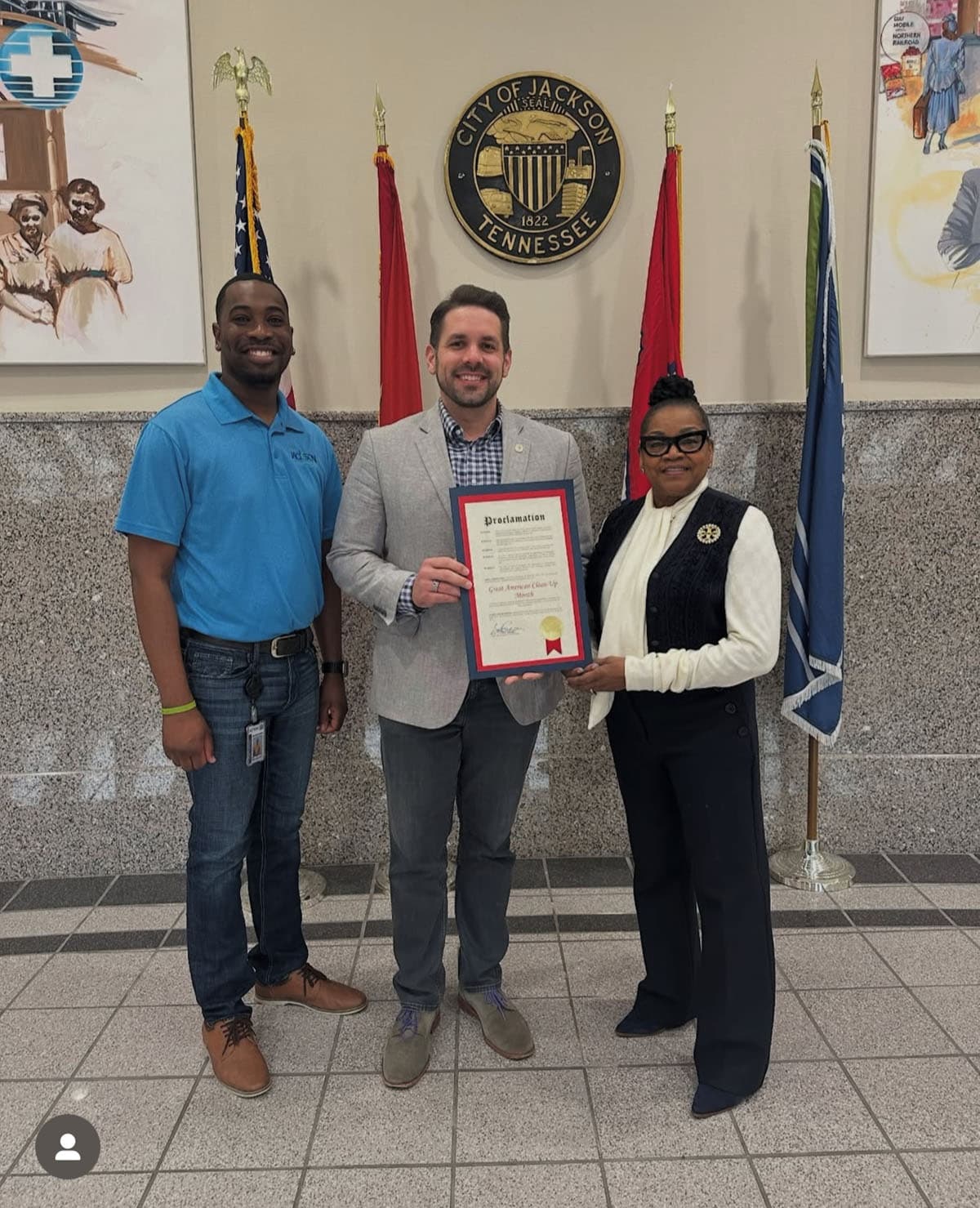Three people hold a framed Proclamation beneath the City of Jackson, Tennessee seal, flanked by American, state and city flags