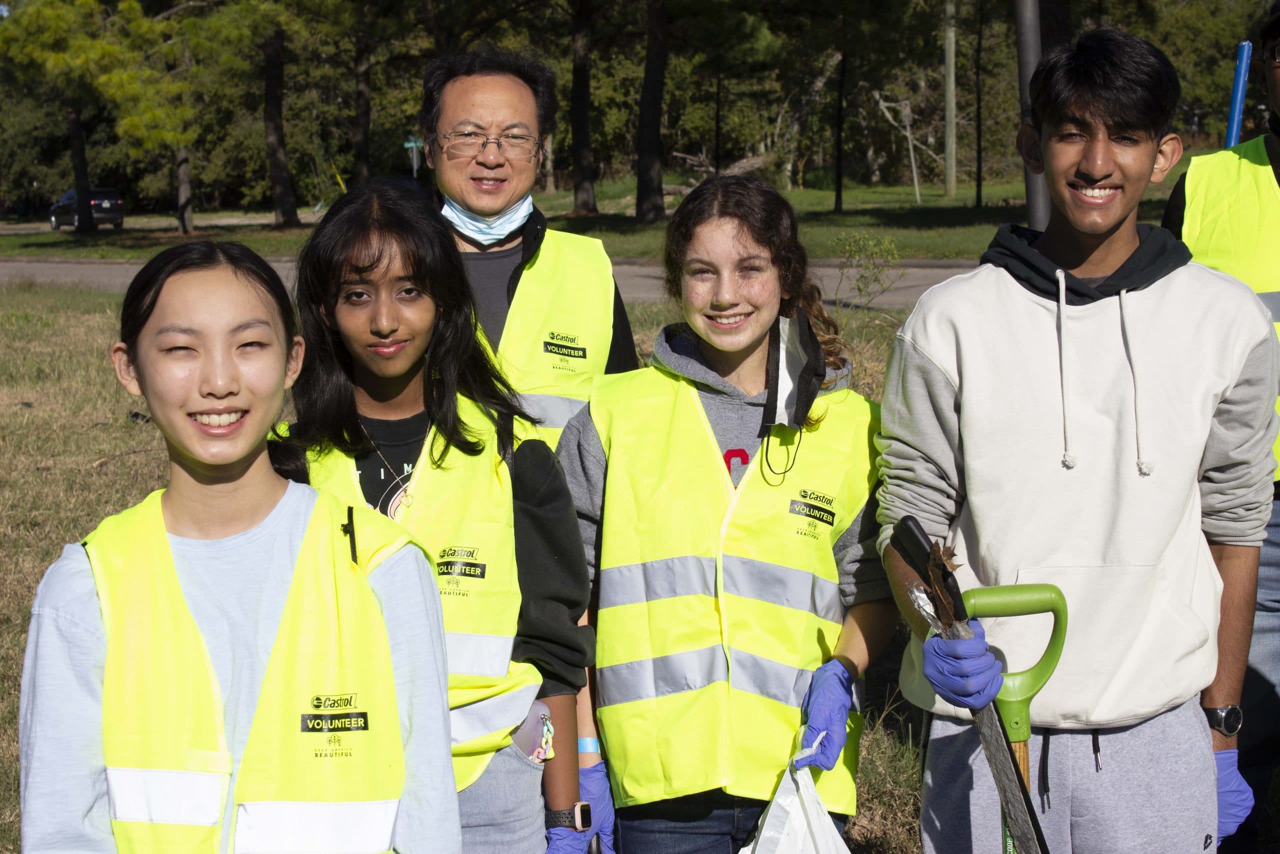 Four students and a masked man in lime Castrol VOLUNTEER vests stand in a park, one holding a shovel and a bag of collected leaves