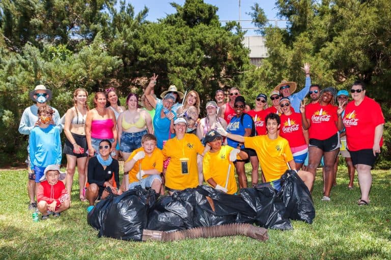 Large mixed group in red Coca-Cola shirts, yellow staff shirts and swimwear pose on grass behind a row of stuffed black trash bags and a dredge pipe