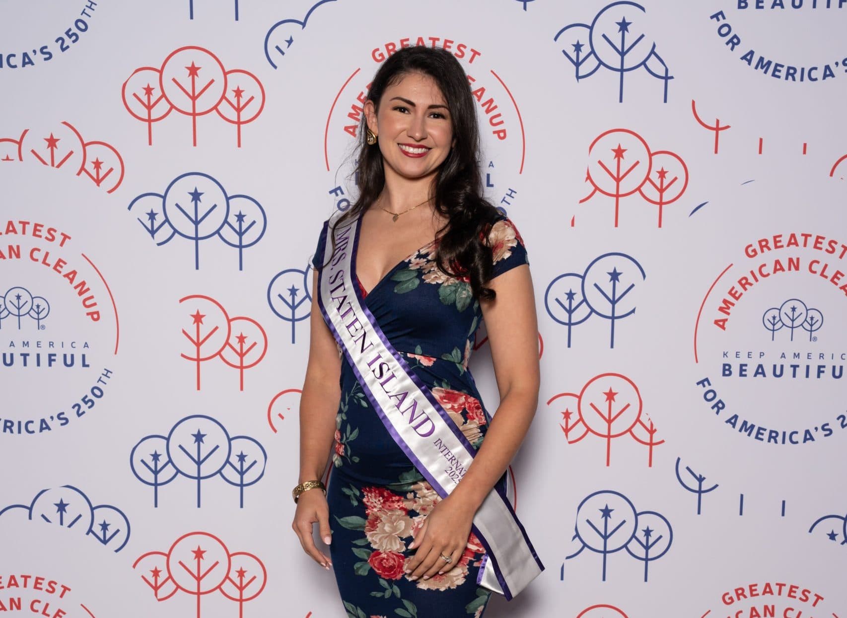 Woman in a navy floral dress wearing a purple-trimmed sash reading Mrs. Staten Island, posed alone at the backdrop