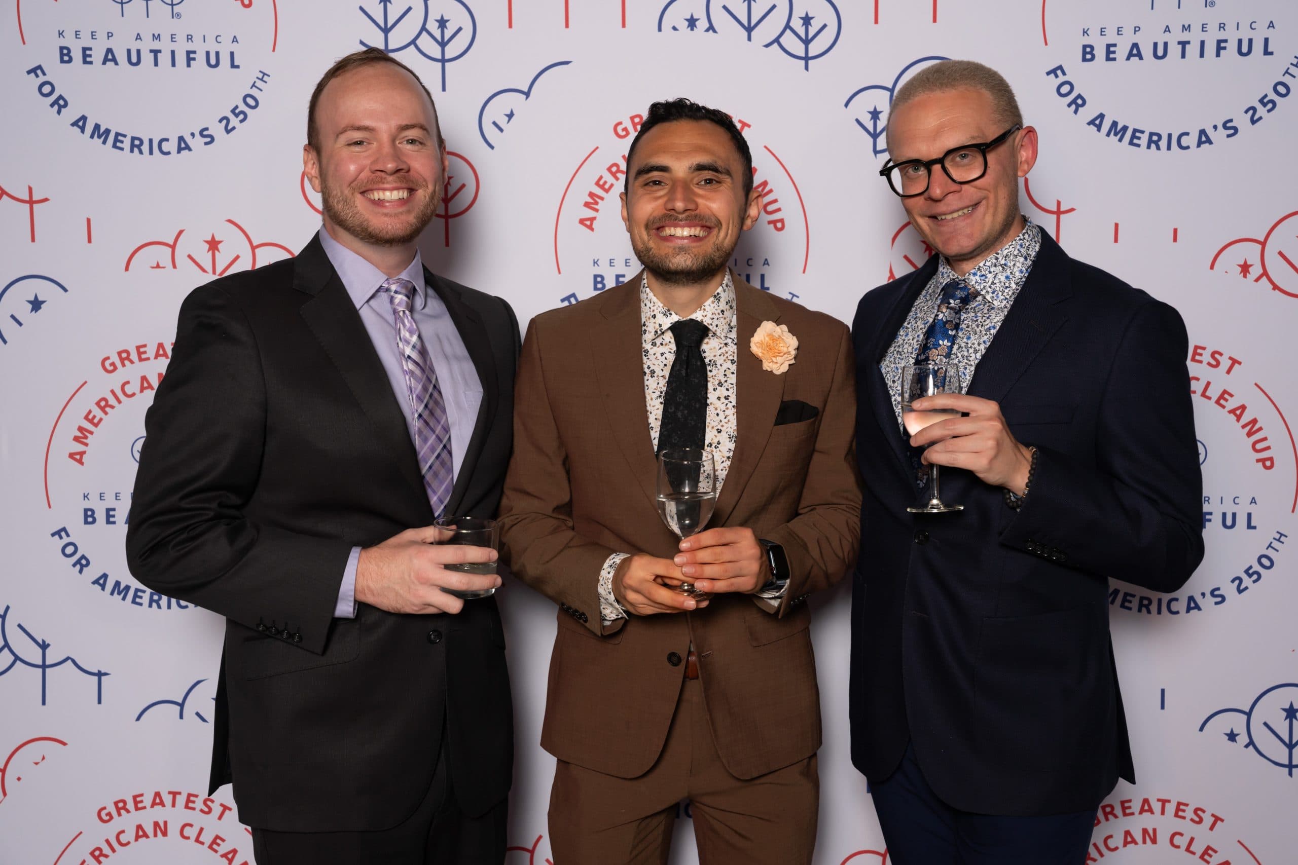 Three men with drinks at the step-and-repeat: one in black with a lilac plaid tie, one in a rust-brown suit with a peach lapel flower, one in navy with a floral shirt