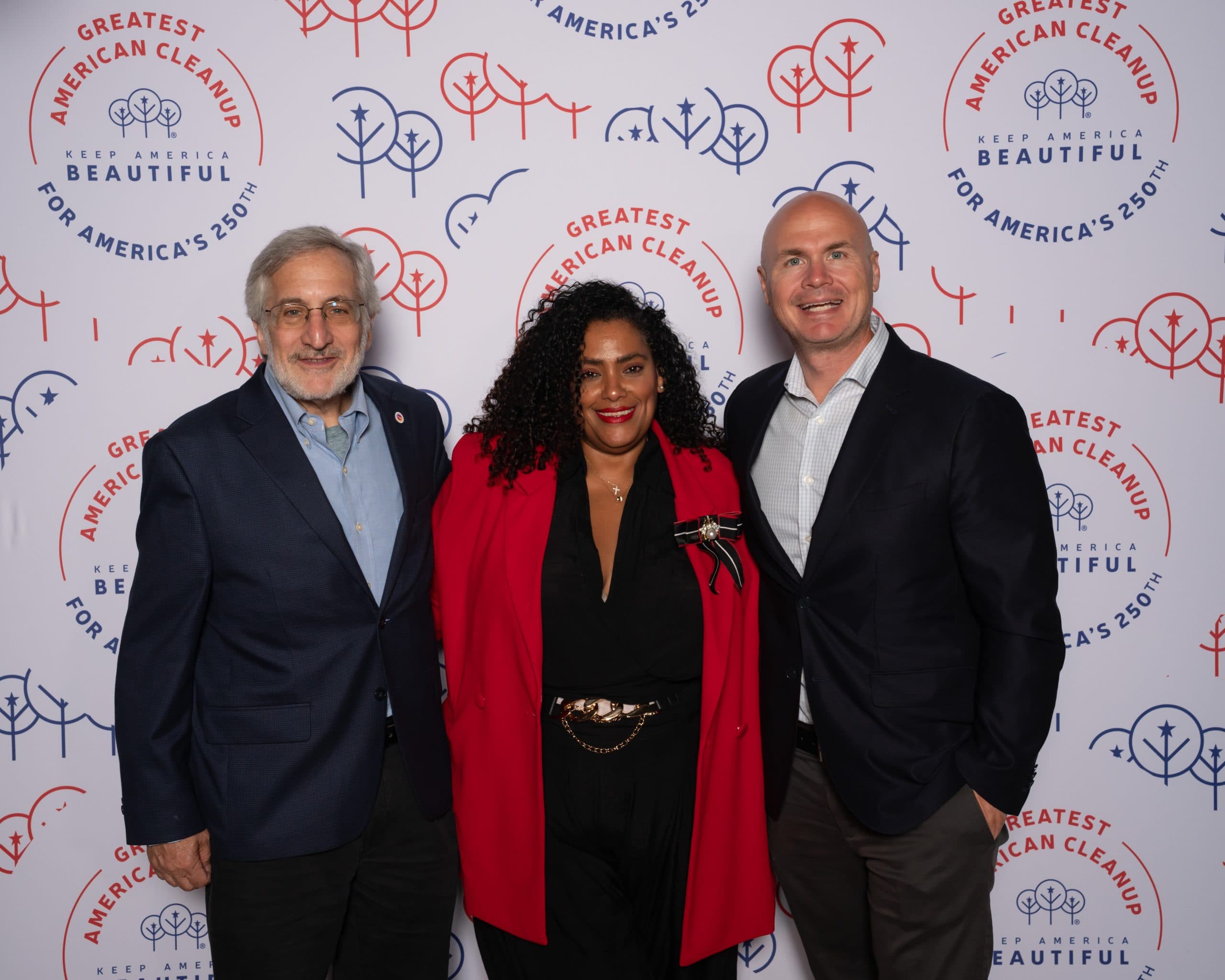 Bearded man in a navy check blazer, a woman in a red blazer over black with a gold chain belt, and a man in a navy jacket and grey trousers