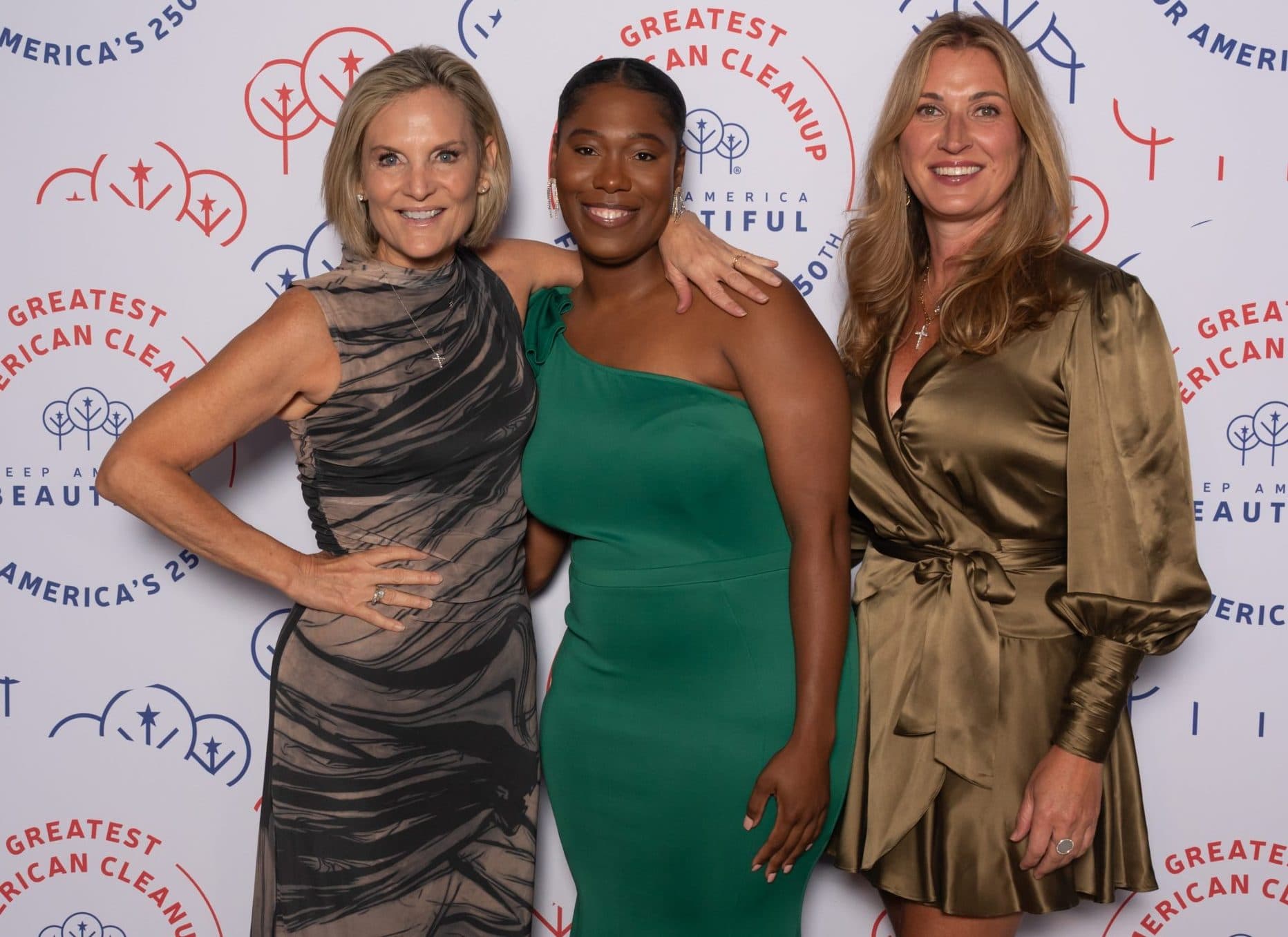 Three women arm in arm: one in a taupe and black print dress, one in an emerald one-shoulder gown, one in a bronze satin wrap dress